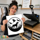 A woman holds up a white square print featuring a black circular duck hunting silhouette design with a dog, three ducks, and weeds. In the background, a black heat press machine sits on a wooden desk next to shelves and clothing racks.