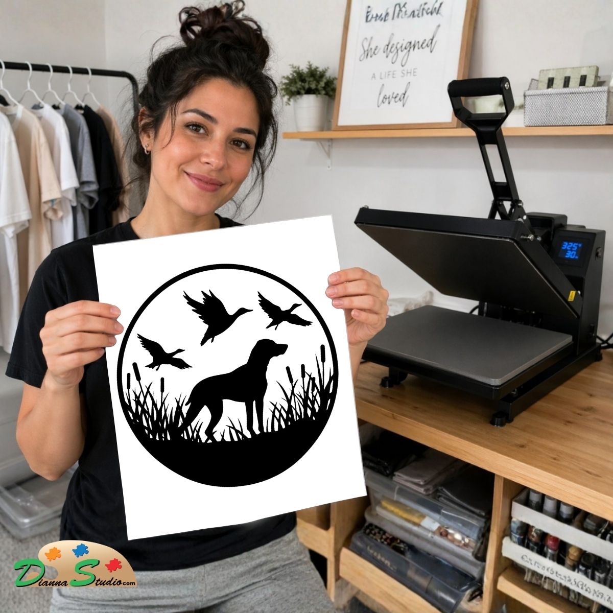 A woman holds up a white square print featuring a black circular duck hunting silhouette design with a dog, three ducks, and weeds. In the background, a black heat press machine sits on a wooden desk next to shelves and clothing racks.