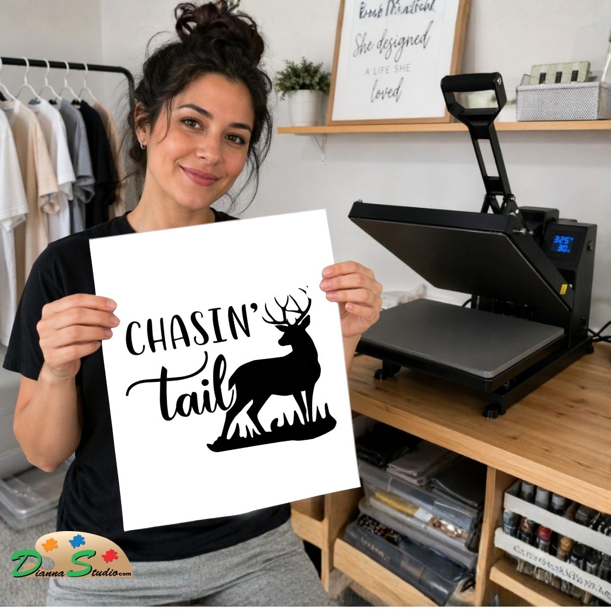 A woman holds up a white print featuring a black deer silhouette and the text 'CHASIN' tail' in a craft room setting with a heat press machine on a wooden desk.