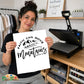 A woman holds up a white print featuring the black text 'FAITH CAN MOVE mountains, MATTHEW 17:20' with a mountain graphic. She is standing in a craft room with a heat press machine on a wooden table behind her.