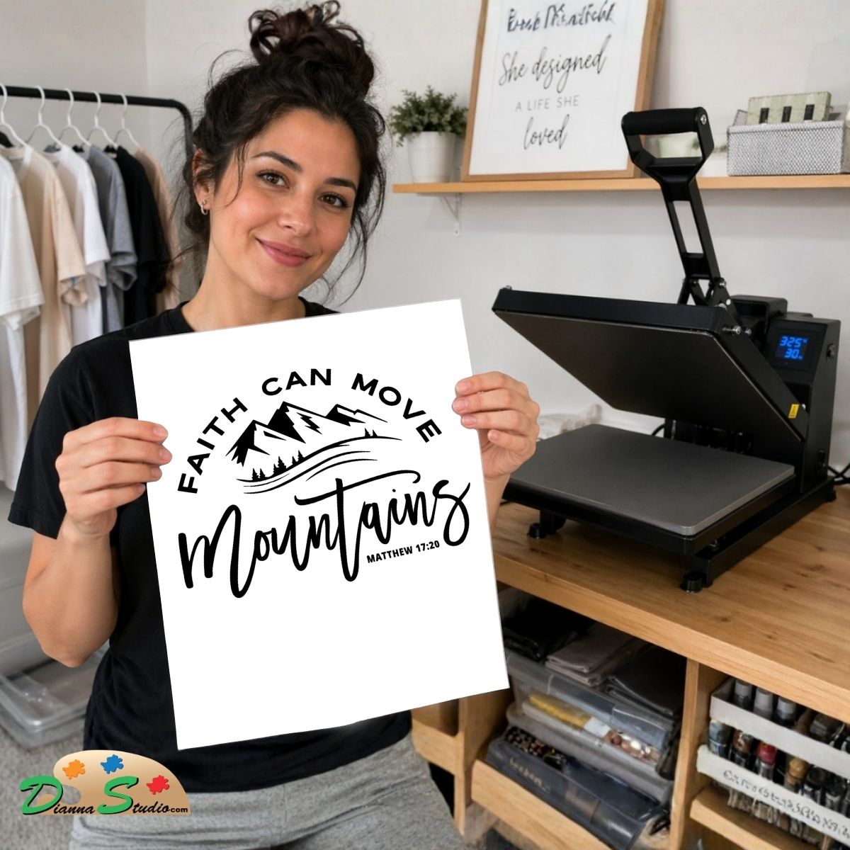 A woman holds up a white print featuring the black text 'FAITH CAN MOVE mountains, MATTHEW 17:20' with a mountain graphic. She is standing in a craft room with a heat press machine on a wooden table behind her.