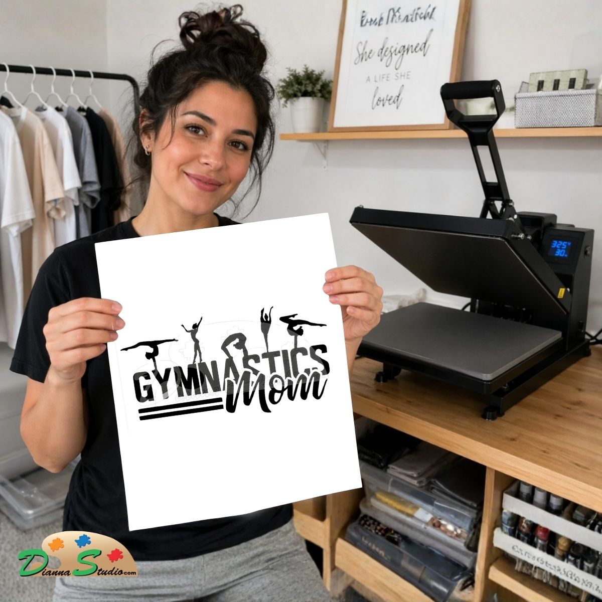 A woman holds up a white graphic print featuring the text 'GYMNASTICS Mom' in black with gymnast silhouettes. She is standing in a home studio environment with a heat press machine on a wooden table behind her.