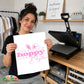 A woman holds up a white square print featuring pink text that reads 'hoppy hoppy Easter' with bunny ears, in front of a wooden craft room setup with a heat press machine.