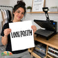 A woman holds up a white print with the text 'LOOK PRETTY' in black  lettering, with 'play dirty' in script written across the center. She is standing in a craft studio with a heat press machine on a wooden table behind her.