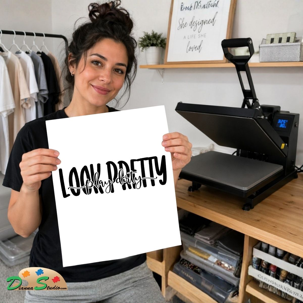 A woman holds up a white print with the text 'LOOK PRETTY' in black  lettering, with 'play dirty' in script written across the center. She is standing in a craft studio with a heat press machine on a wooden table behind her.
