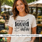 A woman wearing a light beige graphic t-shirt with the words 'loved mom' printed in black. The background is a blurred outdoor patio setting.
