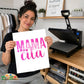 A woman holds up a white square print featuring the text 'MAMA cita' in hot pink  lettering. She is standing in a craft room with a heat press machine on a wooden table behind her.