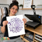 A woman holds up a white square print featuring a purple floral sunburst design, with a heat press machine visible in a craft room setting behind her. Mental Healt Awareness