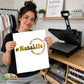 A woman holds up a white heat transfer print featuring the text '#NanaLife' in gold foil-style lettering with the phrases 'LOVE THEM Spoiled' and 'GIVE THEM Back' in smaller curved text. She is standing in a craft room with a black heat press machine on a wooden table behind her.