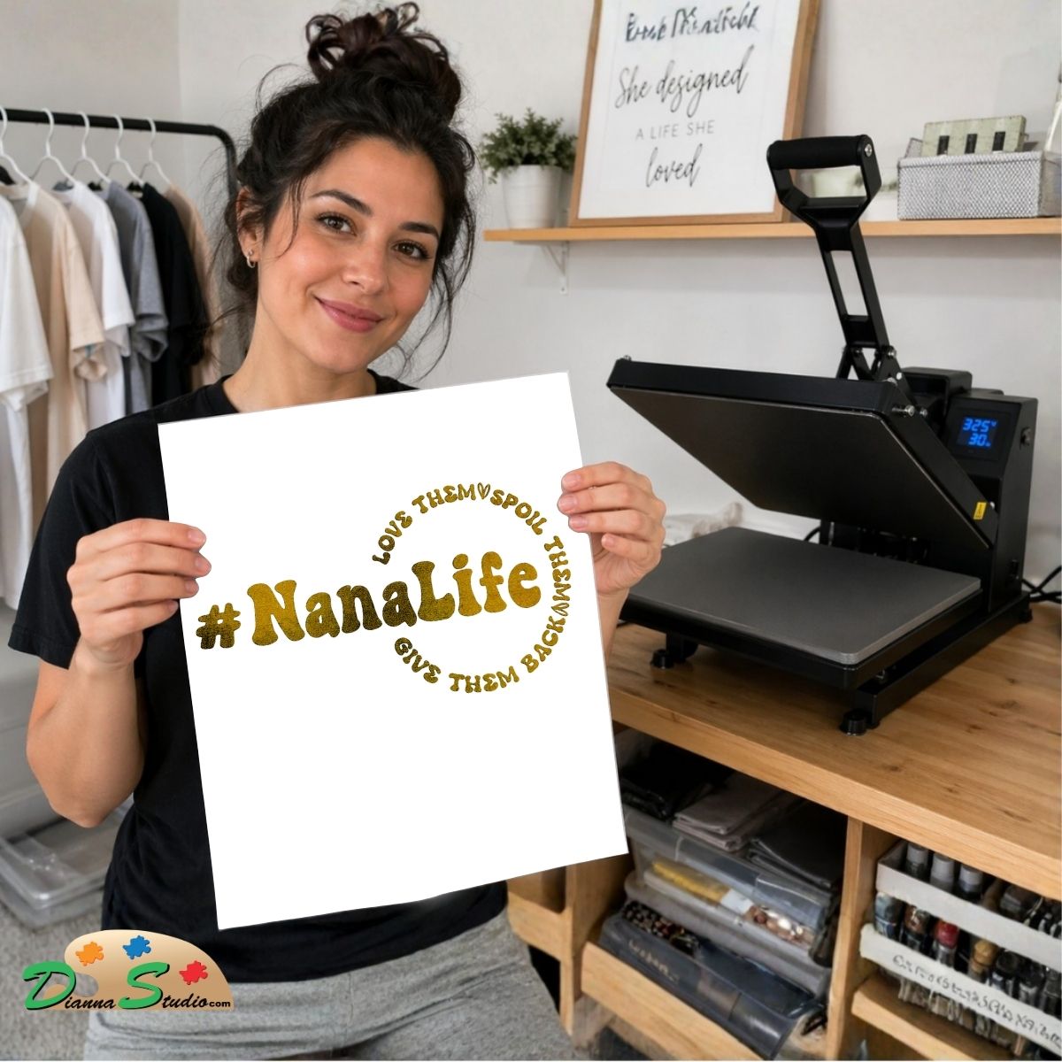 A woman holds up a white heat transfer print featuring the text '#NanaLife' in gold foil-style lettering with the phrases 'LOVE THEM Spoiled' and 'GIVE THEM Back' in smaller curved text. She is standing in a craft room with a black heat press machine on a wooden table behind her.
