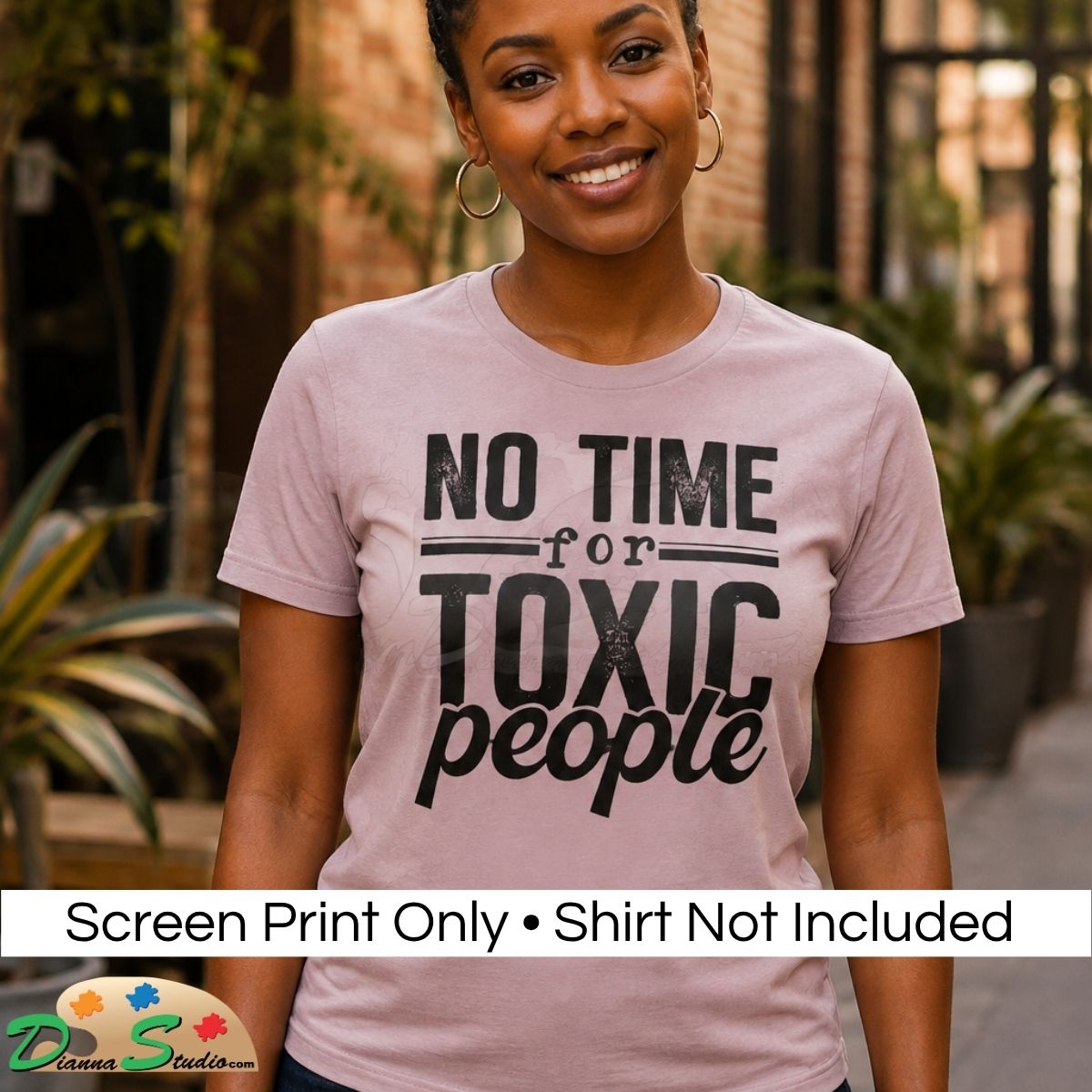 A smiling woman wearing a light pink graphic T-shirt with the text 'NO TIME for TOXIC people' in black. The photo is taken outdoors with blurred plants and a brick wall in the background.