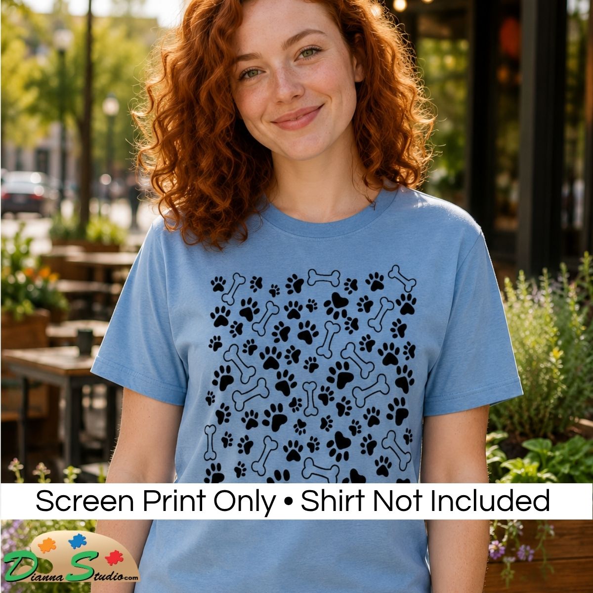 A woman with curly red hair wearing a light blue t-shirt with a black graphic of dog paw prints and bones, set against an outdoor patio background. A text banner at the bottom reads 'Screen Print Only • Shirt Not Included' with a 'DiannaStudio logo.