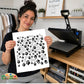 A woman holds up a white sheet featuring black dog-themed silhouettes like bones, hearts, and paw prints, with a heat press machine on a wooden desk in the background.