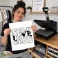 A woman holds up a white square print with the phrase 'SELF LOVE CLUB' in bold black lettering, with a heart symbol replacing the 'O'. She is standing in a craft room with a heat press machine on a wooden table behind her.