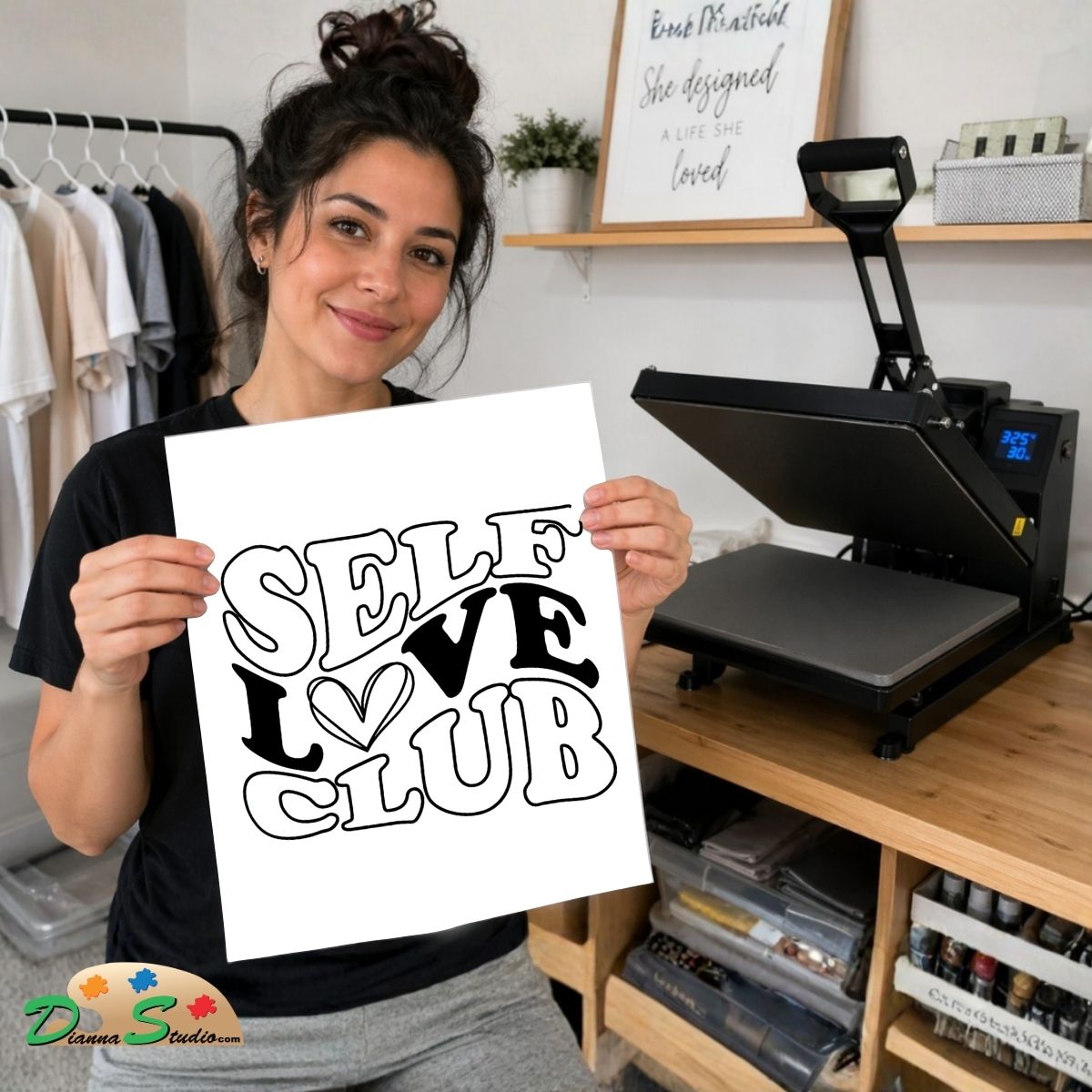 A woman holds up a white square print with the phrase 'SELF LOVE CLUB' in bold black lettering, with a heart symbol replacing the 'O'. She is standing in a craft room with a heat press machine on a wooden table behind her.