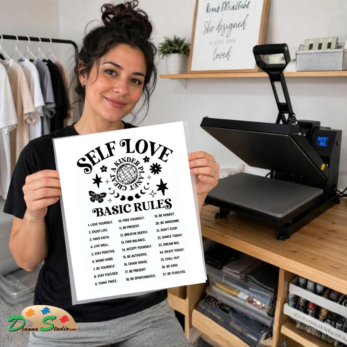 A woman holds up a white heat-transfer sheet with black text reading 'SELF LOVE IS NARROW' and 'BASIC RULES' alongside a checklist. Behind her is a hot press heat press machine, clothing racks, and shelves in a craft room setting.