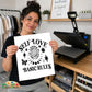 A woman holds up a white square print with black text reading 'SELF LOVE BASIC RULE' and "Create a kinder planet" decorative icons, standing in a craft room with a heat press machine on a wooden table behind her.