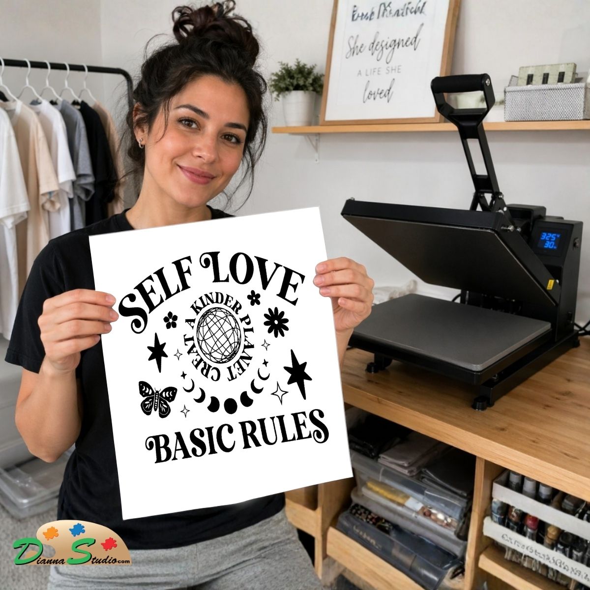 A woman holds up a white square print with black text reading 'SELF LOVE BASIC RULE' and "Create a kinder planet" decorative icons, standing in a craft room with a heat press machine on a wooden table behind her.