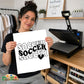 A woman holds up a white print featuring the word 'SOCCER' repeated three times in black retro-style lettering with a black heart underneath. She is standing in a craft room with a heat press machine on a wooden table behind her.
