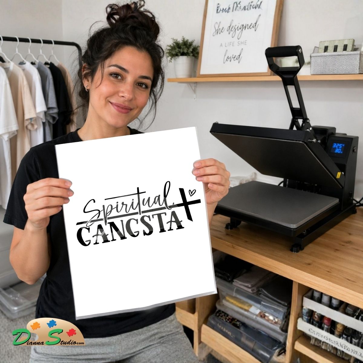 A woman holds up a white square print featuring the black text 'Spiritual GANGSTA' in a casual indoor room with a heat press machine on a wooden table behind her.