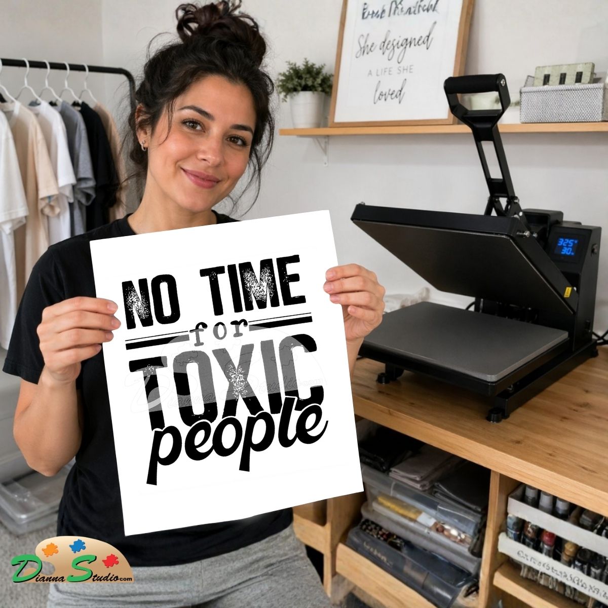 A woman holds up a white square print with the text 'NO TIME for TOXIC people' in black lettering. She is standing in a craft room with a heat press machine on a wooden table behind her.