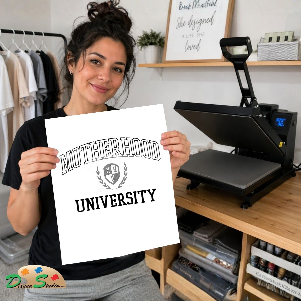 A woman holds up a white print with the text 'MOTHERHOOD UNIVERSITY' and a crest logo. She is standing in a craft room with a heat press machine on a wooden table behind her.