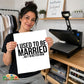 A woman holds up a white square print with bold black text reading 'I USED TO BE MARRIED next to her heat press.