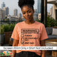 A woman wearing a peach-colored t-shirt with a black graphic print that reads 'WARNING: PLEASE DON'T TRY ME TODAY ON YOUR BEST DAY ANY DAY' with a checked box. The background shows a blurred city skyline and outdoor patio setting.
