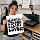A woman holds up a white square print with black text reading 'you are BEAUTIFUL STRONG EARTHY LOVELY' in a craft room setting. A heat press machine sits on a wooden desk behind her, with clothing racks and framed wall art visible in the background.