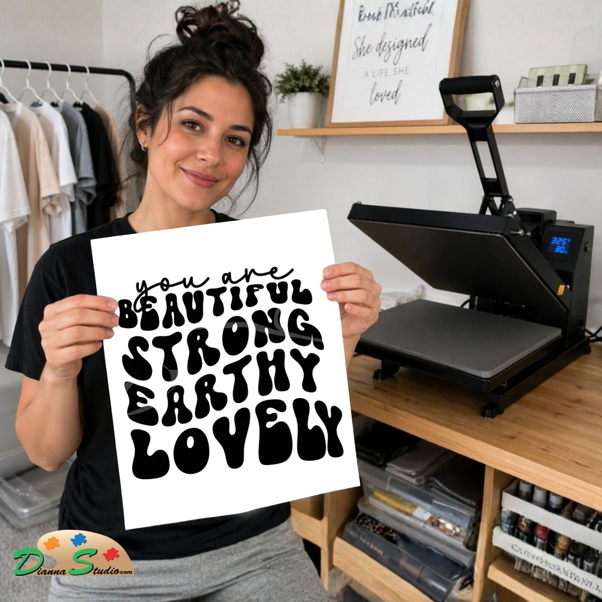 A woman holds up a white square print with black text reading 'you are BEAUTIFUL STRONG EARTHY LOVELY' in a craft room setting. A heat press machine sits on a wooden desk behind her, with clothing racks and framed wall art visible in the background.
